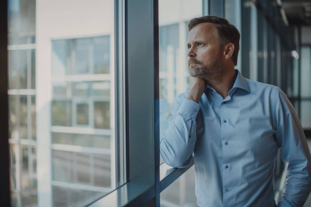 Homme dans la quarantaine regardant par une fenêtre de bureau, moment de réflexion sur sa reconversion professionnelle